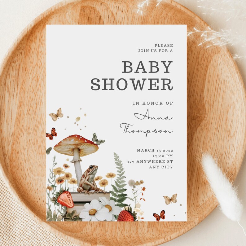May include: A baby shower invitation with a whimsical woodland theme. The invitation features a red and white mushroom, a frog sitting on a stack of books, and butterflies. The text on the invitation reads "Please join us for a baby shower in honor of Anna Thompson, March 13, 2022, 12:00 PM, 123 Anywhere St, Any City."