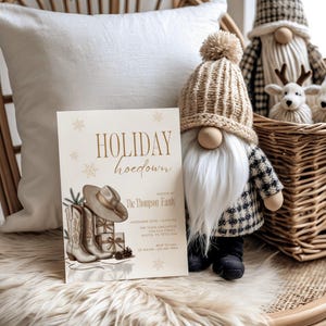 May include: A holiday-themed invitation with the text "HOLIDAY hoedown" and illustrations of cowboy boots, a hat, and wrapped gifts. A gnome wearing a knitted hat and plaid jacket sits next to the invitation. A basket with decorative reindeer is in the background.