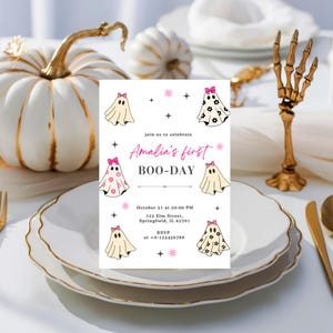 May include: A Halloween-themed invitation with the text "Amalia's first BOO-DAY" and ghost illustrations. The invitation is on a white plate with gold trim, with white pumpkins and a gold skeleton hand in the background.