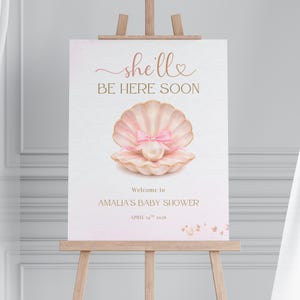 May include: A baby shower welcome sign on an easel. The sign features the text "she'll BE HERE SOON" in pink script, along with a watercolor illustration of a seashell with a pearl and bow. The sign also includes the text "Welcome to AMALIA'S BABY SHOWER" and the date.