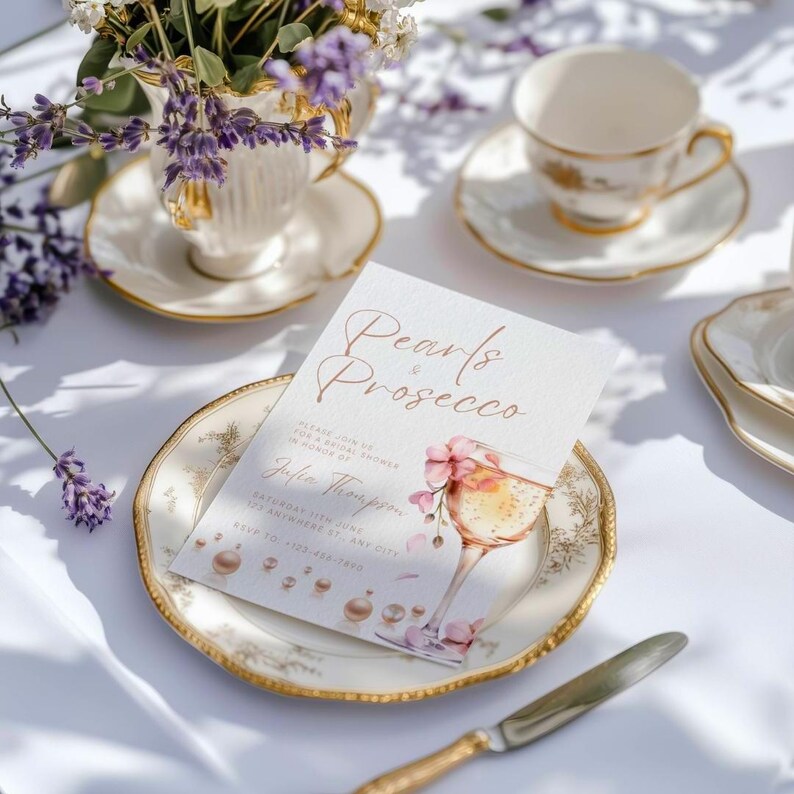 May include: A white card with gold lettering that reads "Pearls & Prosecco" with a watercolor illustration of a glass of champagne and pink flowers. The card is on a white plate with a gold rim.