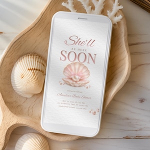 May include: A smartphone displays a baby shower invitation with the text "She'll Be Here Soon." The design features a pink seashell with a pearl, surrounded by smaller pearls. The phone rests on a wooden tray with seashells.