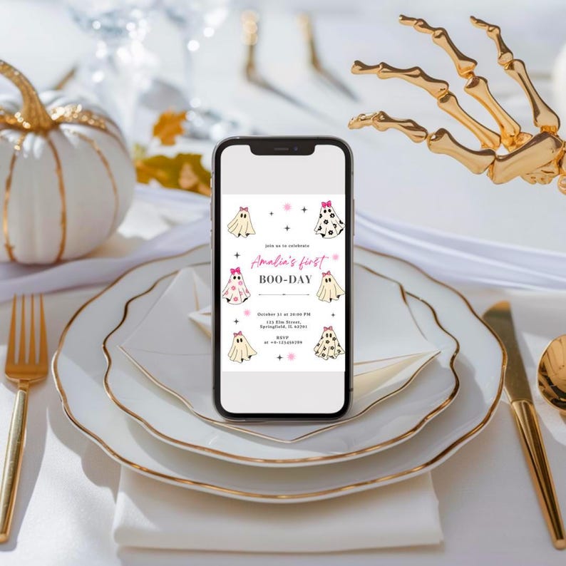 May include: A smartphone displays a Halloween-themed invitation with ghost illustrations and the text "Amelia's first BOO-DAY." The invitation is set on a white plate with gold trim, alongside gold cutlery and a decorative pumpkin. A golden skeleton hand is in the background.