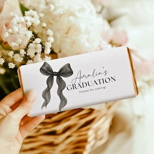 Op de afbeelding: Een witte chocoladereepverpakking met een zwarte aquarelboog en de tekst "Amalia's GRADUATION Thanks for coming!". De verpakking heeft gouden randen en wordt door een persoon vastgehouden.