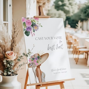 May include: A white sign with a floral design and the text "Cast your vote for best hat" with a drawing of a woman wearing a hat with flowers on it. The sign also says "[Name]'s Bridal Shower, Savannah, Georgia".