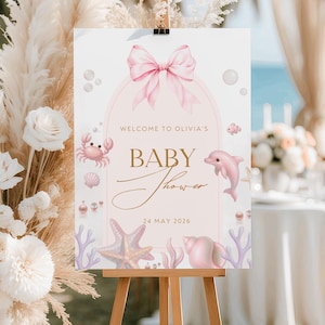 Puede incluir: Un letrero de bienvenida para baby shower sobre un caballete. El letrero presenta un lazo rosa, las palabras "Welcome to Olivia's" y "Baby Shower" en escritura dorada. Ilustraciones de un cangrejo, delfín, estrella de mar y conchas decoran el letrero.