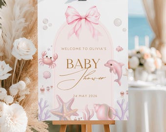 May include: A baby shower welcome sign on an easel. The sign features a pink bow, the words "Welcome to Olivia's", and "Baby Shower" in gold script. Illustrations of a crab, dolphin, starfish, and seashells decorate the sign.
