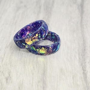 Puede incluir: Dos anillos de resina púrpura con purpurina iridiscente incrustada en la resina. Los anillos están apilados uno encima del otro.