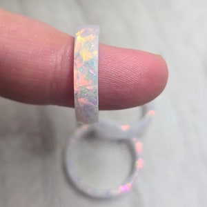 以下が含まれることがあります： 樹脂に埋め込まれた虹色のグリッターが付いた白いリング。リングは指につけられています。