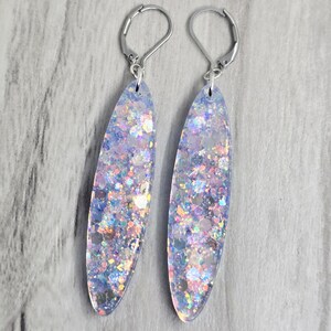 May include: A pair of dangle earrings with a clear, oval design filled with iridescent glitter. The earrings have silver lever-back closures. The glitter reflects pink, blue, and silver. The earrings are approximately 6 cm long.