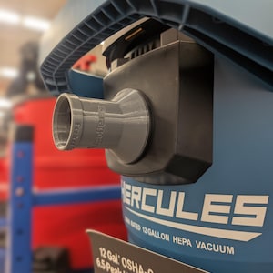 Puede incluir: Primer plano de una aspiradora HEPA Hercules azul y negra de 12 galones. La aspiradora tiene una boquilla gris y un asa. El texto "Hercules" es visible en el lateral. Una etiqueta indica que cumple con la OSHA.