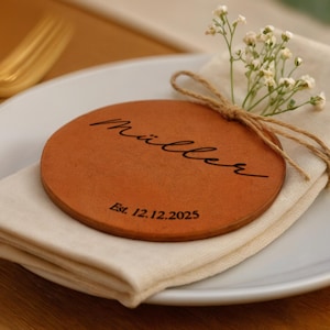 Könnte beinhalten: Eine runde, braune Platzkarte aus Leder mit dem Namen "Müller" und dem Datum "Est. 12.12.2025" in schwarzer Schrift. Die Platzkarte liegt auf einer weißen Serviette, die mit Bindfaden gebunden und mit kleinen weißen Blumen verziert ist. Eine goldene Gabel und ein Messer sind sichtbar.