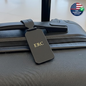 Könnte beinhalten: Ein schwarzes Gepäckanhänger mit den Initialen "ERC" in goldfarbenen Buchstaben hängt am Griff eines schwarzen Koffers. Das Etikett ist rechteckig mit abgerundeten Ecken. Der Koffer hat eine strukturierte Oberfläche und einen Reißverschluss.