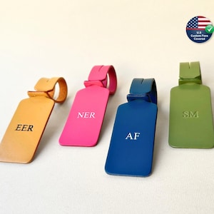 May include: Four colorful luggage tags in yellow, pink, blue, and green. Each tag has a different set of initials: EER, NER, AF, and SM. The tags are made of a smooth material and have a loop for attaching to luggage. A U.S. Custom Fees Covered sticker is in the background.