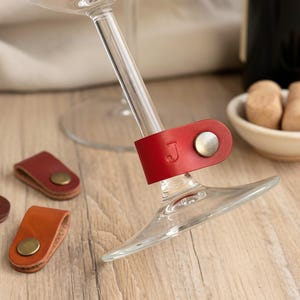 Puede incluir: Un conjunto de colgantes de cuero de colores para copas de vino. Un colgante rojo con la letra "J" está sujeto a una copa de vino. Otros colgantes son verdes, amarillos, marrones, naranjas y morados. Cada colgante tiene un broche metálico.