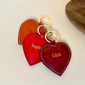 Puede incluir: Tres llaveros de cuero en forma de corazón en color tostado, rojo brillante y rojo oscuro. Cada llavero tiene costuras blancas en contraste, una anilla de metal dorado y texto personalizado: 'MC' en el tostado, 'Sophie' en el rojo brillante y 'E&N' en el rojo oscuro.