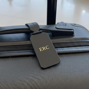 Könnte beinhalten: Schwarzes Gepäckanhänger mit den Initialen "ERC" in goldfarbenen Buchstaben. Das Etikett ist an einem schwarzen Koffer mit Griff befestigt. Der Koffer hat eine strukturierte Oberfläche und einen Reißverschluss.