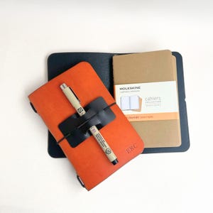 Może przedstawiać: Pomarańczowy skórzany notatnik z czarnym uchwytem na długopis i dwoma długopisami. Notatnik Moleskine Cahiers Collection znajduje się obok notatnika. Notatnik ma inicjały "ERC" w prawym dolnym rogu.