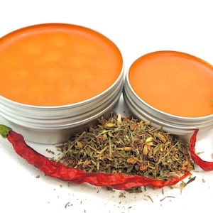 May include: Two round, silver tins filled with orange balm, alongside dried herbs and red chili peppers. The balm has a smooth, glossy surface. The herbs are a mix of green and brown. The chili peppers are a vibrant red.