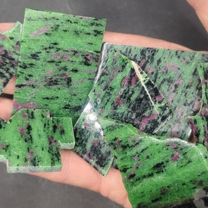 May include: A collection of green, black, and pink gemstone rough. The rough is in various shapes and sizes.
