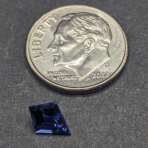 May include: A dark blue faceted gemstone with a kite shape. The gemstone is sitting on a gray surface next to a 2022 United States dime.