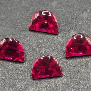 May include: Four semi-circular, faceted, ruby-red gemstones. The gems are arranged on a dark gray surface. Each gem has a polished, glossy appearance, reflecting light. The facets create a sparkling effect, enhancing the vibrant color.