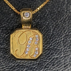 May include: A gold pendant necklace with a square-shaped charm featuring a cursive letter 'A' with small diamonds set around it.
