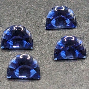 May include: Four blue gemstone cabochons, each shaped like a half-circle, are arranged on a gray surface. The gemstones are smooth and polished, with a deep blue color.