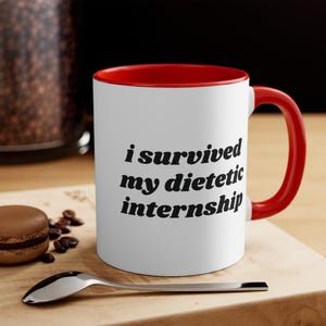May include: White ceramic coffee mug with a red rim and handle. The mug has black text that reads "i survived my dietetic internship".