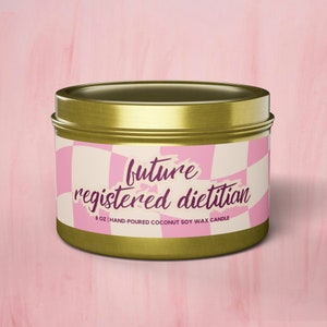 Puede incluir: Lata de vela dorada con un patrón de cuadros blancos y rosas y el texto "future registered dietitian" y "8 oz | HAND-POURED COCONUT SOY WAX CANDLE".