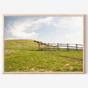Peut inclure: Une clôture en bois avec un portail menant à une colline herbeuse avec un ciel bleu et des nuages blancs en arrière-plan.