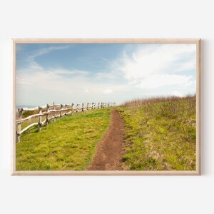 Peut inclure: Un chemin de terre serpente à travers un champ herbeux, bordé d'une clôture en bois. Le ciel est bleu avec des nuages blancs.