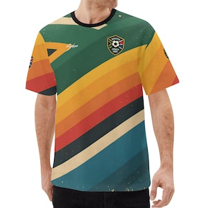 K&ouml;nnte beinhalten: Ein Fu&szlig;balltrikot im Retro-Stil mit einem Design aus gr&uuml;nen, gelben, orangefarbenen, roten und blauen Streifen. Das Shirt hat einen schwarzen Kragen und das Emblem S&uuml;dafrika 1991 auf der Brust. Das Trikot hat einen Vintage-Look.