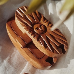 Könnte beinhalten: Eine Holzbox mit einem geschnitzten Sonnendesign auf dem Deckel. Die Box ist geöffnet und gibt den Innenraum frei, und ein kleines Holzstück liegt in der Nähe. Das Holz hat einen warmen, rötlich-braunen Ton, und das Gesicht der Sonne ist detailliert.