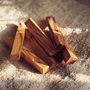 May include: A group of five wooden sticks, likely Palo Santo, arranged on a beige textured surface. The sticks are light brown with darker brown streaks and are slightly curved.