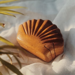 May include: A wooden box in the shape of a seashell, with a hinged lid. The box is made of light brown wood and has a carved, ribbed design on the top. The box is resting on a white cloth.