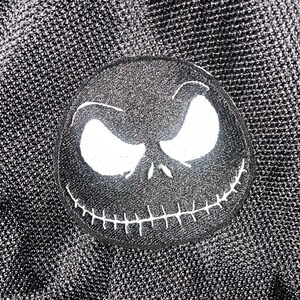 May include: Black and white embroidered patch of Jack Skellington's face from the Nightmare Before Christmas film. The patch has a stitched smile and white eyes.