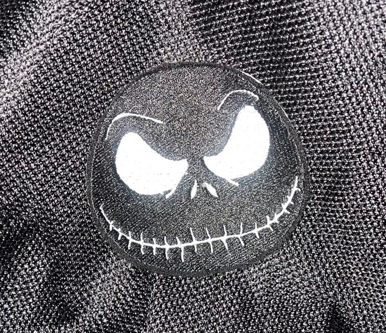 May include: Black and white embroidered patch of Jack Skellington's face from the Nightmare Before Christmas film. The patch has a stitched smile and white eyes.