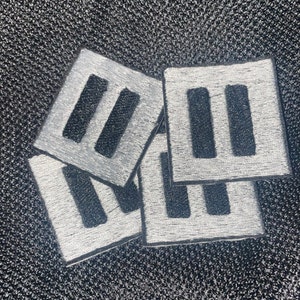 May include: Four silver-coloured fabric patches with black embroidered stripes. Each patch is square-shaped and has three parallel stripes.