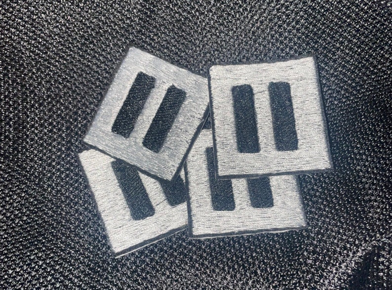 May include: Four silver-coloured fabric patches with black embroidered stripes. Each patch is square-shaped and has three parallel stripes.
