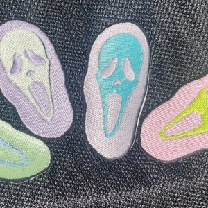 Könnte beinhalten: Vier gestickte Patches in verschiedenen Pastellfarben, jede mit einem weißen Geistergesicht mit einem Schrei. Die Patches befinden sich auf einem schwarzen Stoffhintergrund.