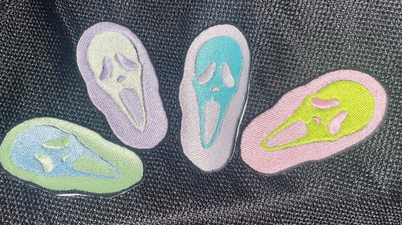 May include: Four embroidered patches in different pastel colours, each featuring a white ghost face with a scream. The patches are on a black fabric background.