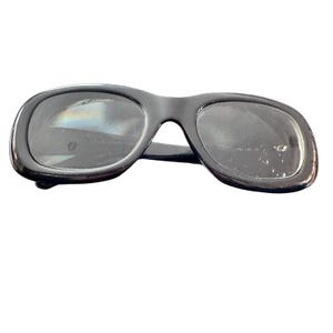 Può includere: Occhiali da sole neri con una montatura spessa e arrotondata. Le lenti sono scure e il ponte è stretto. Gli occhiali da sole sono su uno sfondo bianco.