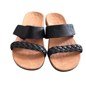 Könnte beinhalten: Ein Paar schwarze Sandalen mit geflochtenem Riemen über den Zehen und einem breiten Riemen über dem Fuß. Das Fußbett ist beige und strukturiert. Die Sandalen sind auf weißem Hintergrund.