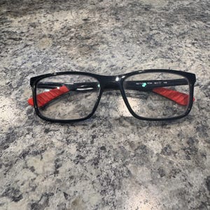 Könnte beinhalten: Schwarze rechteckige Brille mit roten Akzenten an den Bügeln. Die Brille hat klare Gläser und einen schwarzen Rahmen. Der Text "RB 7124 54 0 17 140" ist auf dem rechten Bügel aufgedruckt.