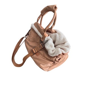 Peut inclure: Un sac de transport pour animaux de compagnie de couleur pêche avec un intérieur moelleux de couleur crème. Le sac est doté d'une bandoulière et de poignées marron. Le sac est doté d'un fermoir en métal et d'une poignée en cuir.