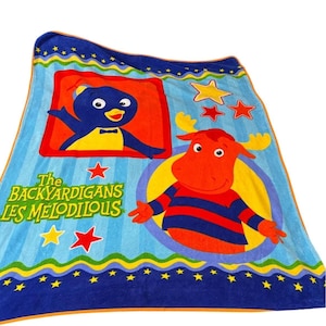 May include: A blue Backyardigans blanket featuring cartoon characters Pablo and Tyrone. The blanket has a blue border with yellow stars and a light blue background with vertical stripes. The text "The Backyardigans Les Mélodilous" is also visible.