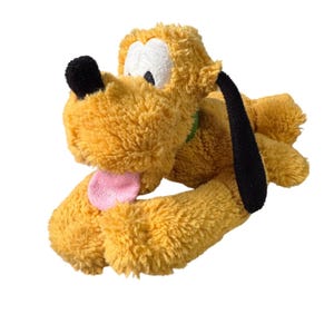 May include: A soft toy of Pluto, a cartoon dog. The toy is mainly yellow with black ears and nose, white eyes, and a pink tongue. It has a green collar and is lying down.