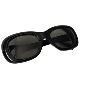 Könnte beinhalten: Schwarze Sonnenbrille mit einem abgerundeten, ovalen Rahmen. Die Gläser sind dunkel und der Rahmen hat eine glänzende Oberfläche. Die Sonnenbrille ist leicht angewinkelt, wodurch die Form der Gläser und die Dicke des Rahmens sichtbar werden.