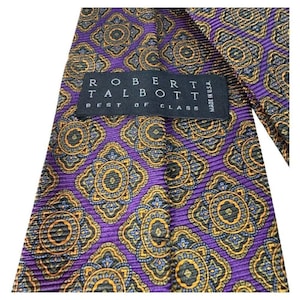 Puede incluir: Corbata Robert Talbott morada con un patrón repetido de medallones florales dorados, azules y negros. La etiqueta dice "ROBERT TALBOTT BEST OF CLASS MADE IN U.S.A."